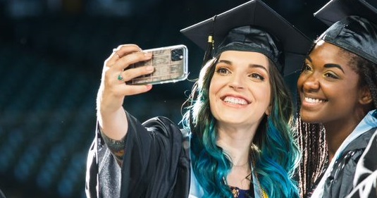  two graduates in caps and gowns taking a selfie with their phones 