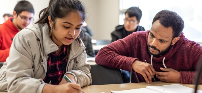 two students in a classroom setting reading from a book