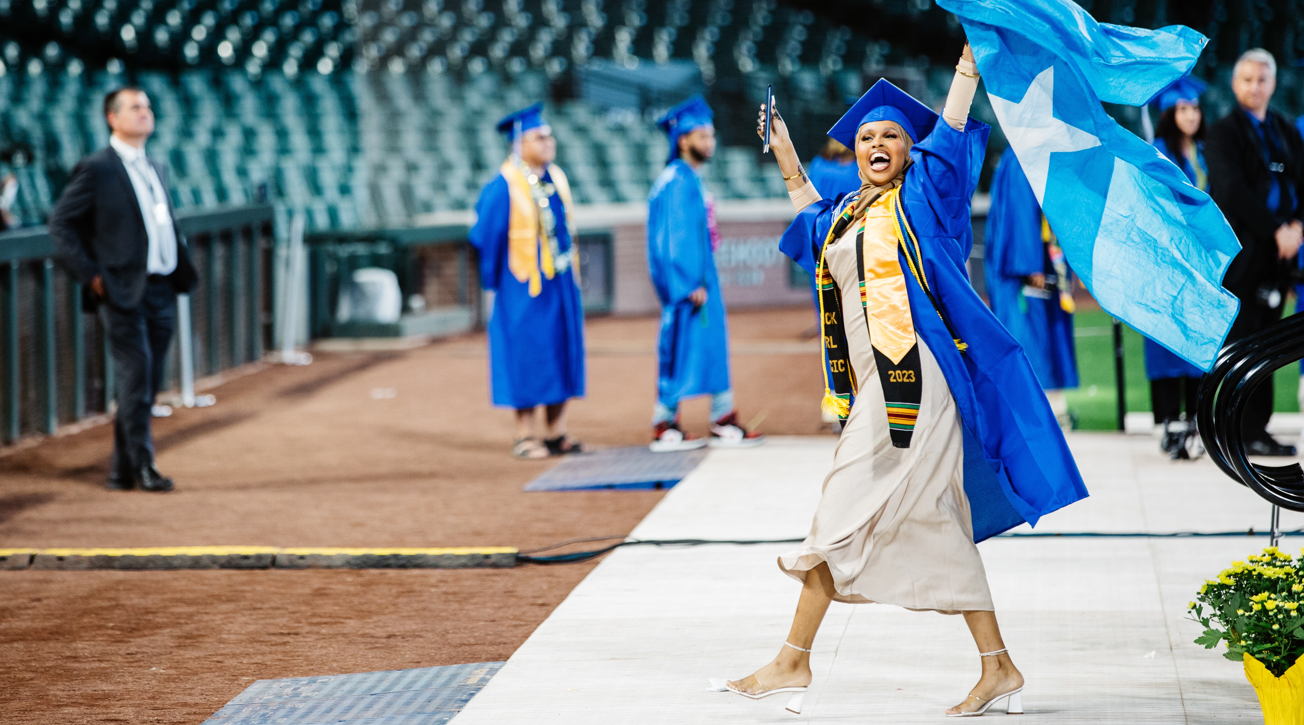  student in a cap and gown walking off the commencement stage smiling and holding a flag 