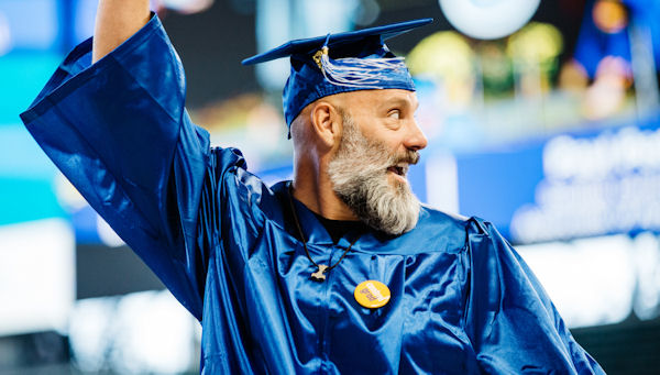  A male graduate with a beard who is smiling and waving 