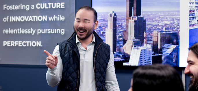 A corporate representative smiling and pointing to a sign that says "Fostering a culture of innovation while relentlessly pursuing perfection."