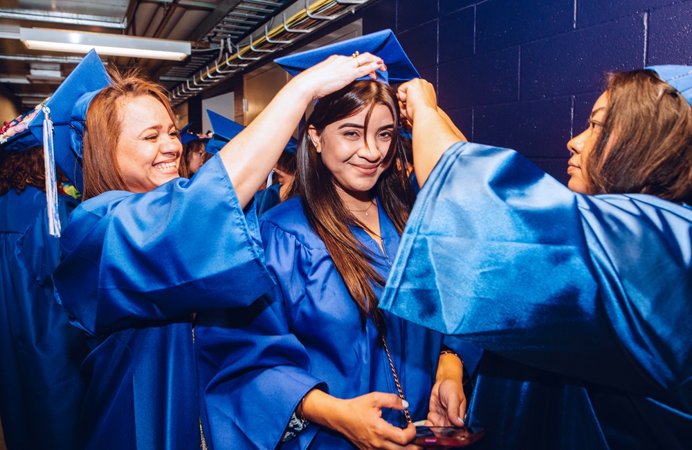 Thee students in blue graduation caps and gowns adjusting their caps.