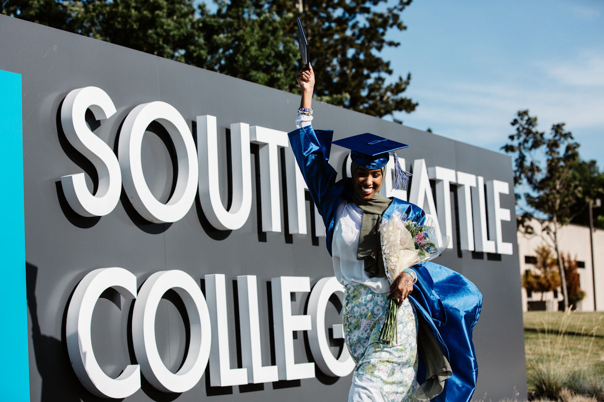 student in cap and gown holding flowers and walking in front of South Seattle College sign