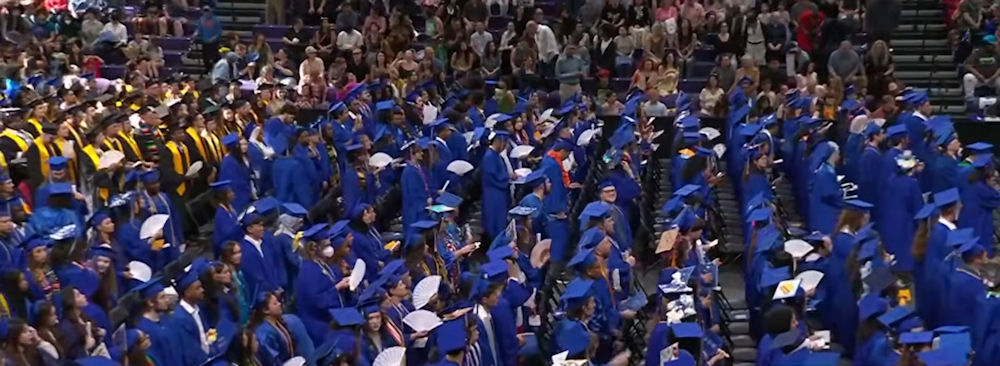 a large group of graduates in caps and gowns on the main floor of the arena during the commencement ceremony