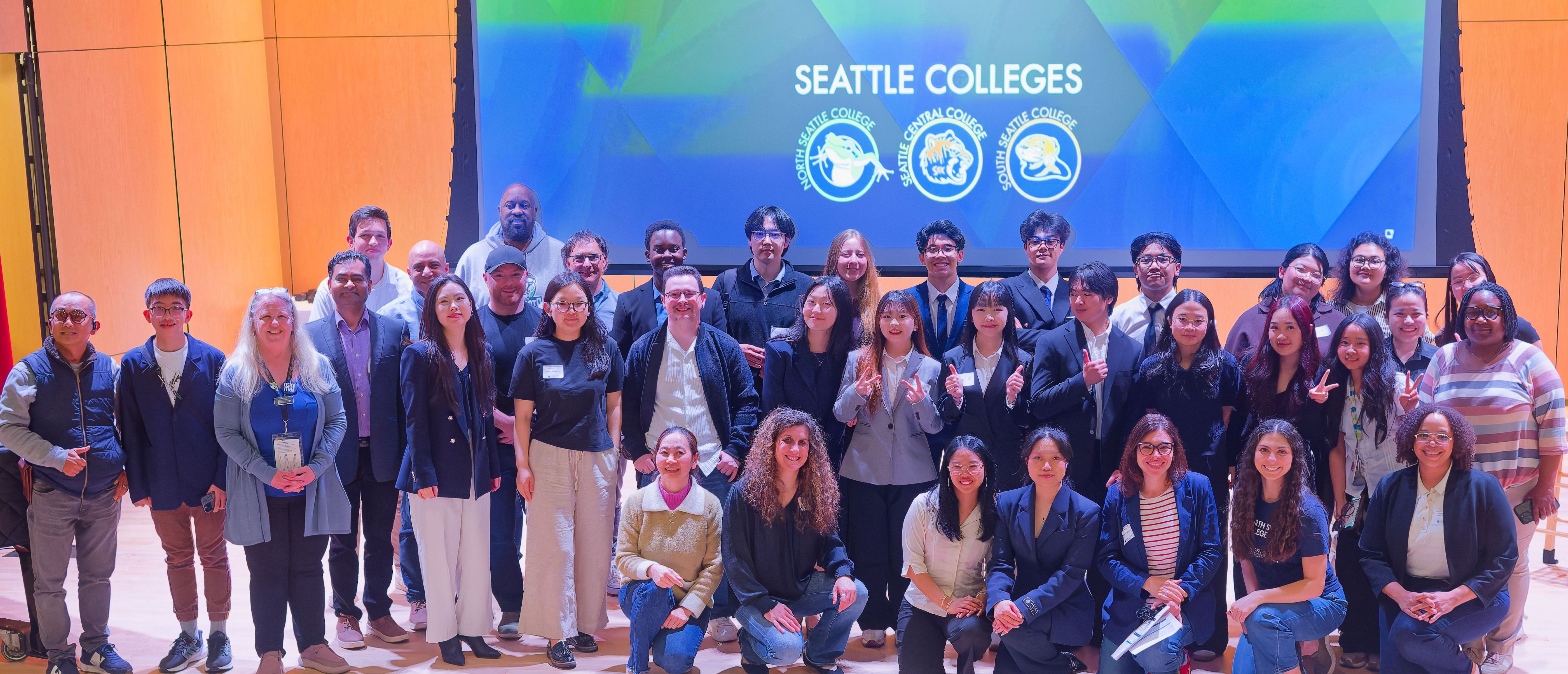 large group of students standing on a stage with blue background on a screen with Seattle Colleges mascots on it